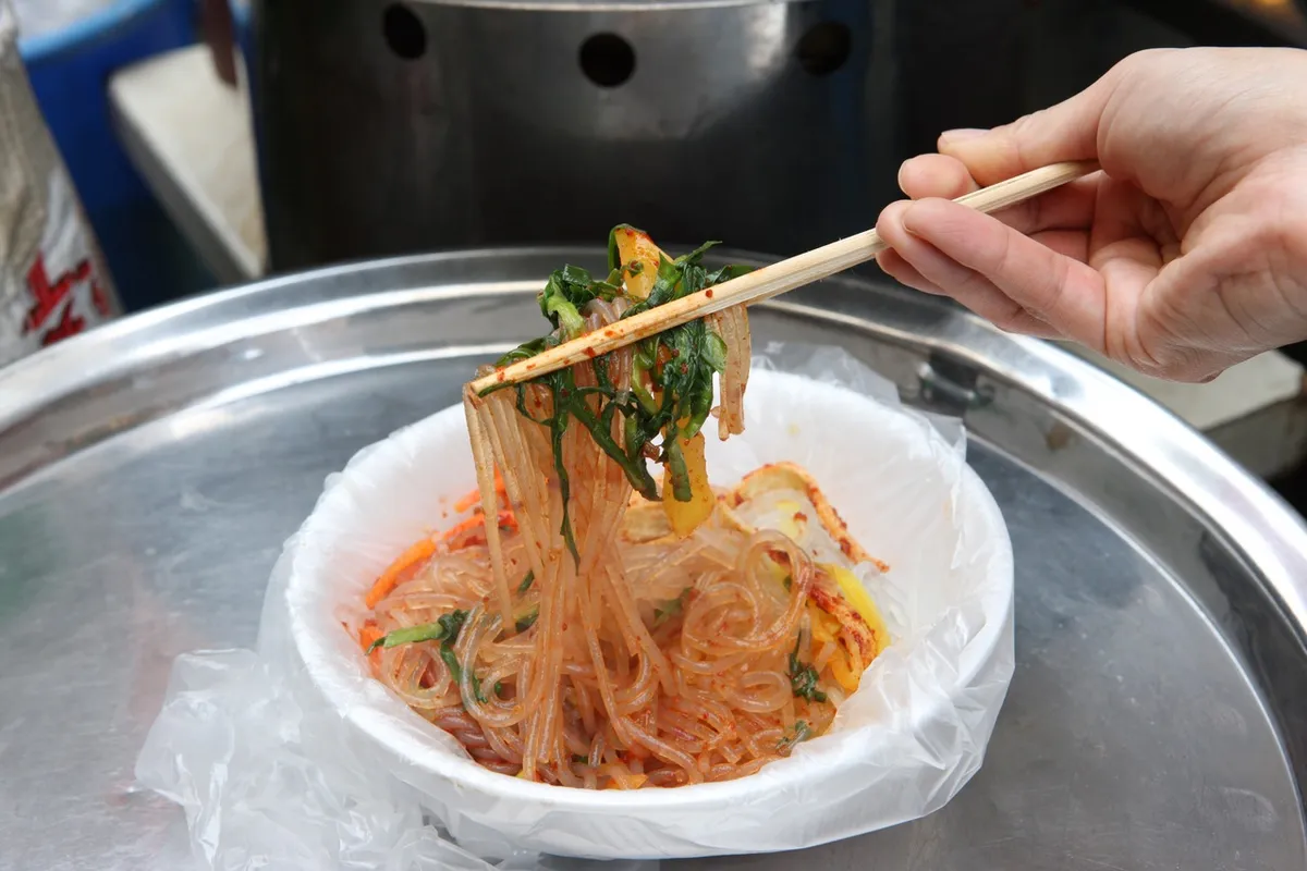 Chopsticks lifting red-seasoned bibim dangmyeon glass noodles in Busan