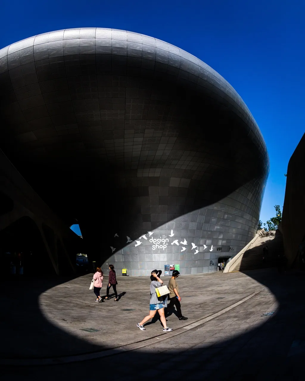 Visitors beneath the curved overhang of Dongdaemun Design Plaza against a blue sky