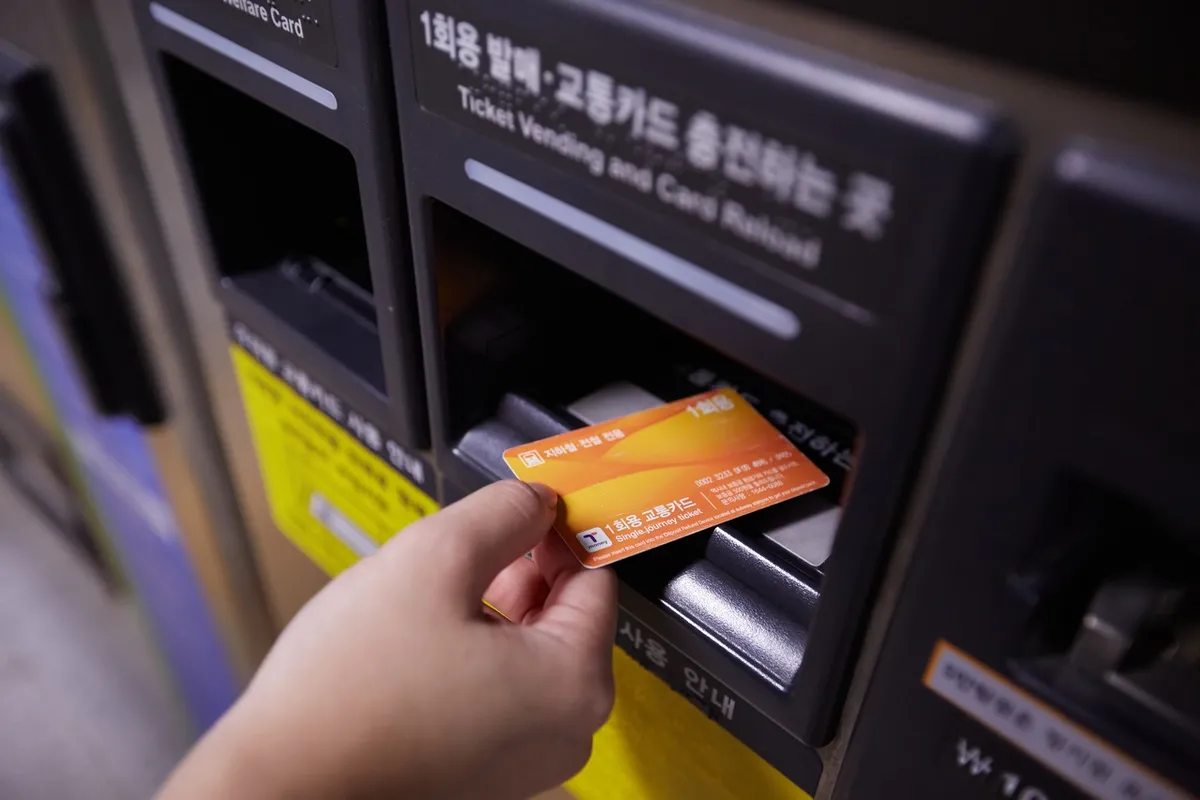 Hand holding an orange single-journey subway card at a ticket machine in Seoul.