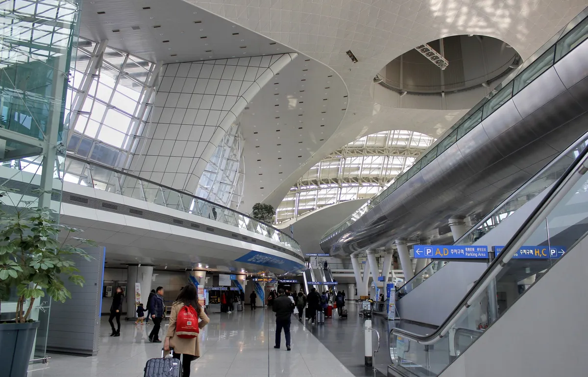 Travelers with luggage and escalators beneath train and parking signs in Incheon Airport Transportation Center