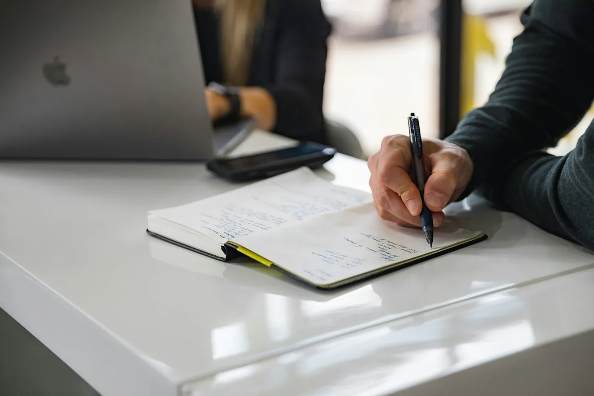 Hand writing notes in an open notebook beside a laptop and smartphone