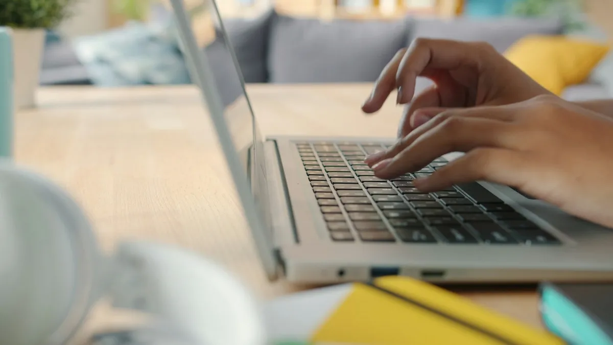 Hands typing on a silver laptop at a wooden table with headphones and a yellow notebook