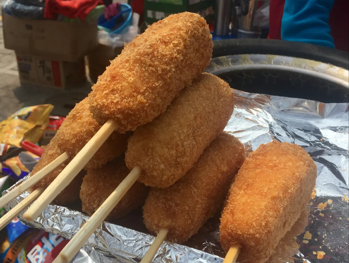Several breadcrumb-coated Korean corn dogs on wooden sticks above a foil-lined food-stall tray