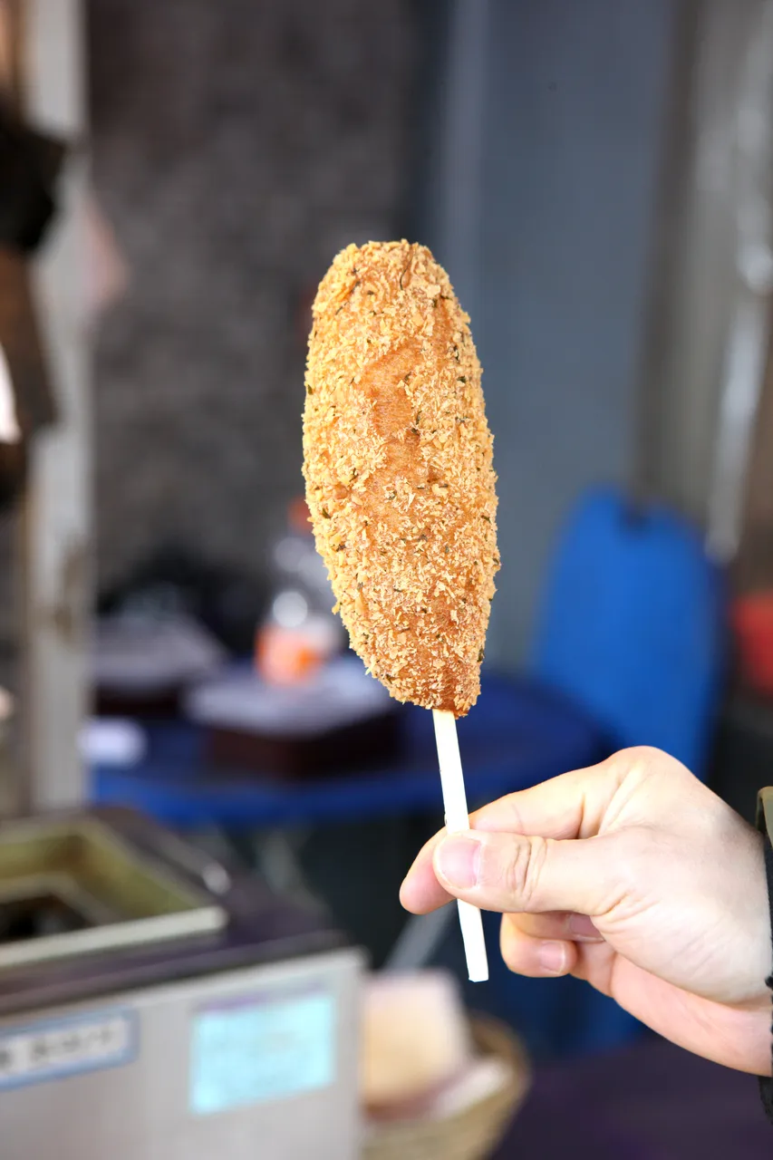 Hand holding a golden breadcrumb-coated Korean corn dog upright on a wooden stick at a food stall