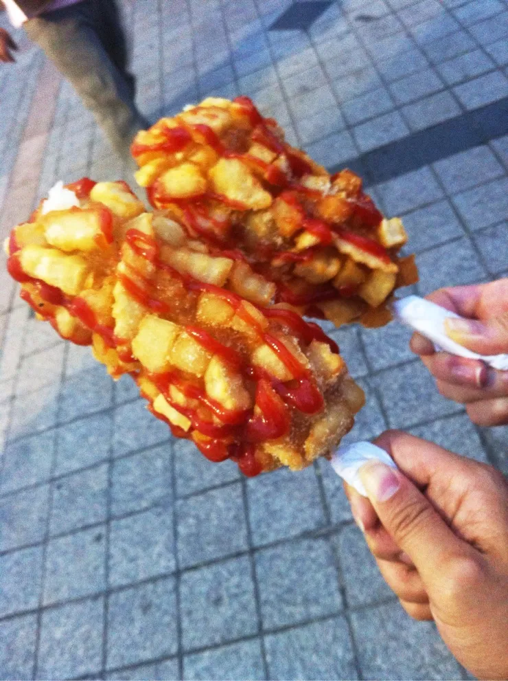 Two hands holding potato-coated Korean corn dogs drizzled with red and yellow sauces over a paved sidewalk