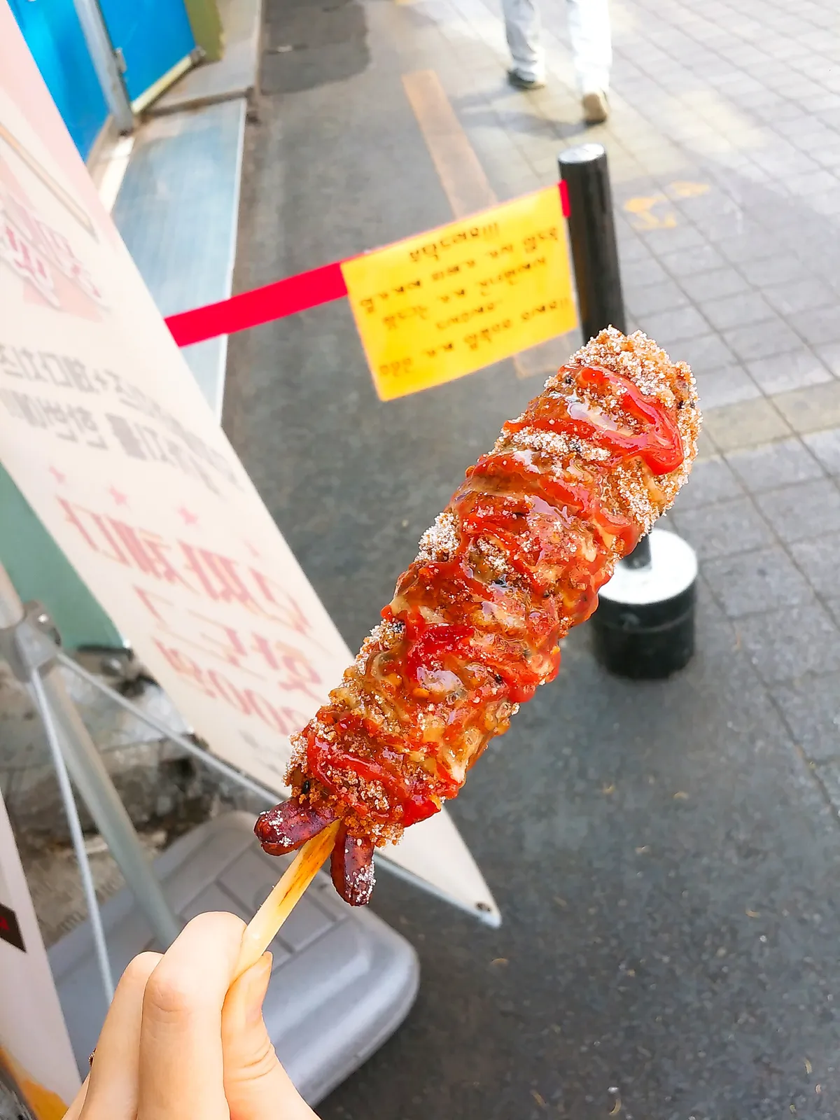 Hand holding a textured Korean corn dog drizzled with red sauce beside a street-food stall