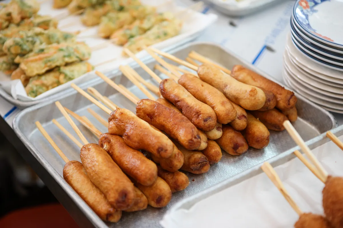 Golden-brown Korean corn dogs on wooden sticks in a stainless-steel market display beside stacked plates