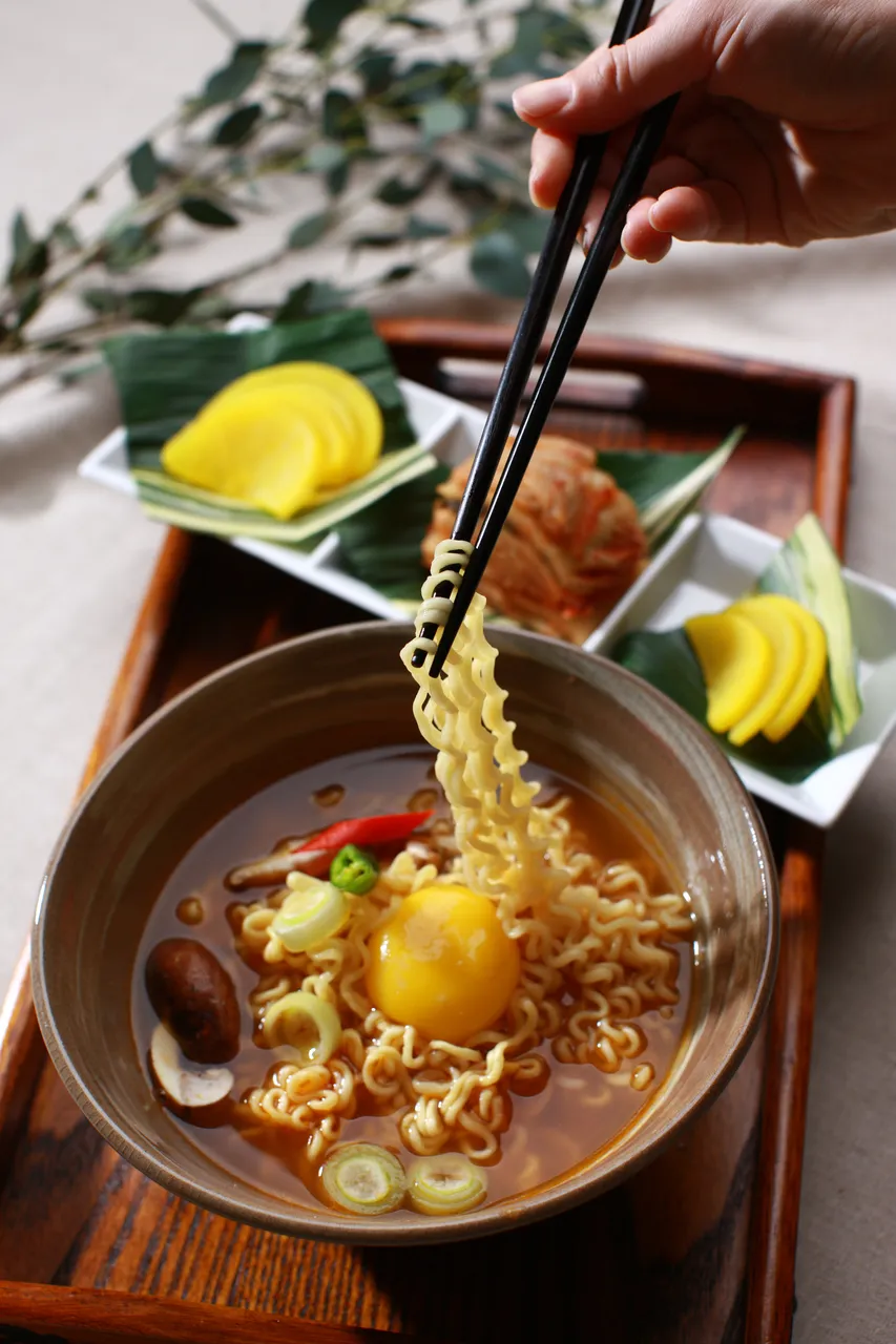 Korean ramyeon noodles lifted with chopsticks beside kimchi and pickled radish