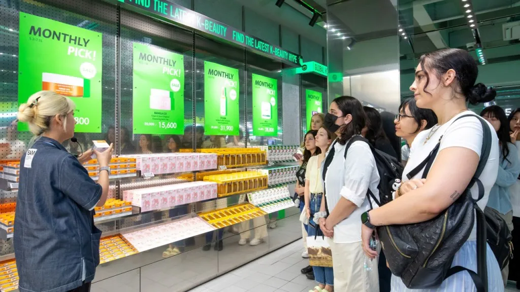 Olive Young staff member addressing shoppers beside stocked cosmetics shelves and green display panels