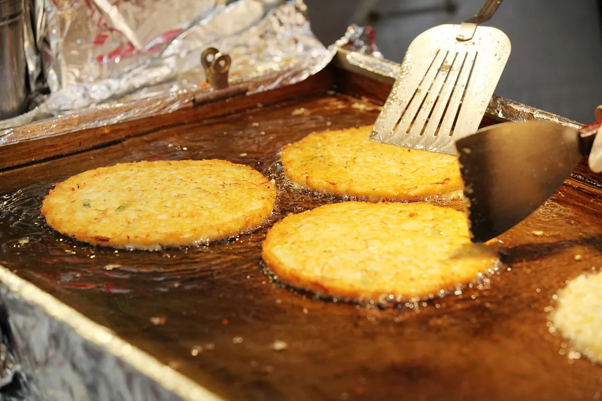 Golden bindaetteok pancakes cooking on a griddle beside metal spatulas at Gwangjang Market in Seoul