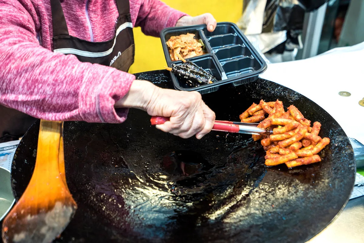 Red-coated rice cakes on a round griddle beside a person holding a lunchbox at Tongin Market in Seoul
