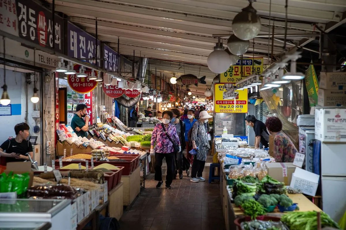 Fresh green vegetables displayed beside shoppers and food stalls in an aisle at Gyeongdong Market in Seoul