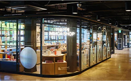 Glass-fronted MMCA Seoul shop with illuminated shelves and merchandise displays beside a museum corridor