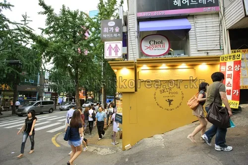 Pedestrians beside a yellow corner storefront and commercial buildings on Yonsei-ro in Sinchon