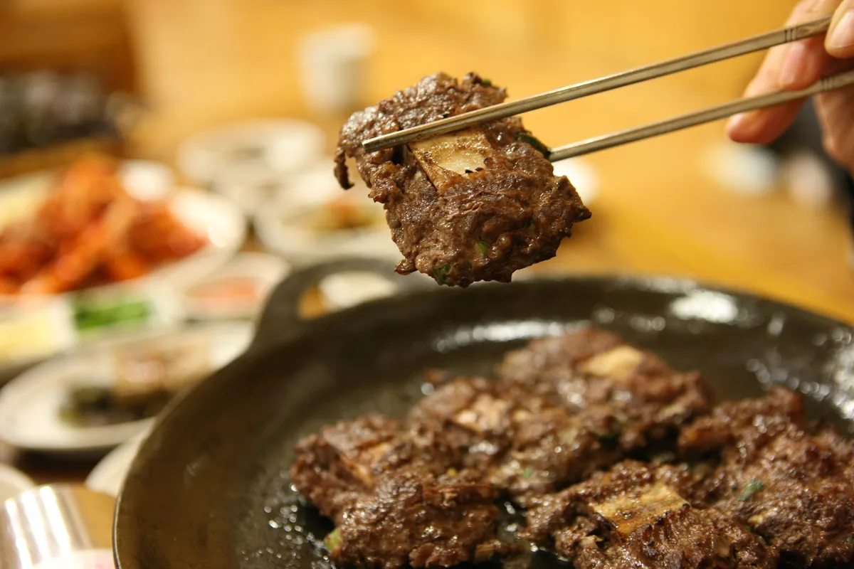 Piece of grilled tteokgalbi lifted with chopsticks above a cooking pan