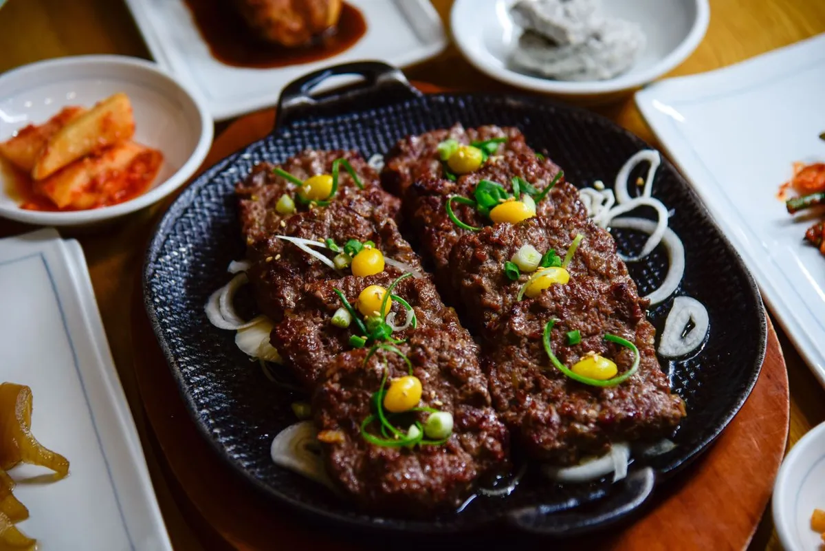 Grilled tteokgalbi patties on a black plate with sliced onions and side dishes